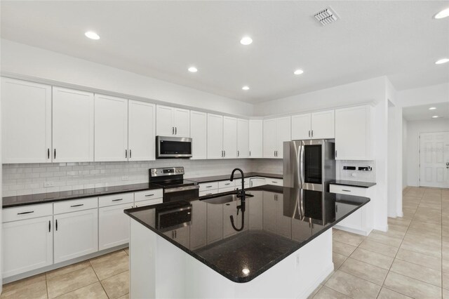 kitchen with an island with sink, sink, white cabinetry, stainless steel appliances, and dark stone counters