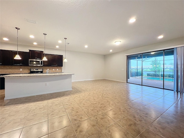 kitchen with decorative backsplash, an island with sink, appliances with stainless steel finishes, hanging light fixtures, and light stone countertops