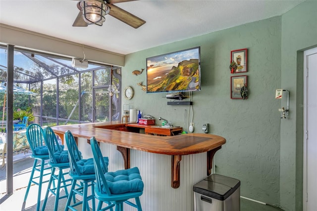 bar featuring a textured wall, a bar, a ceiling fan, and a sunroom