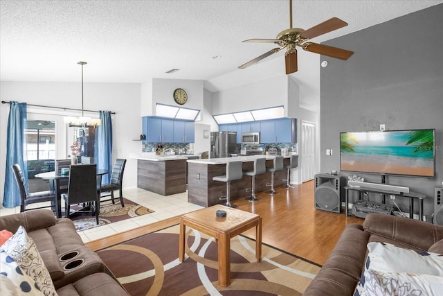 living room featuring high vaulted ceiling, light wood-style flooring, a textured ceiling, and ceiling fan with notable chandelier