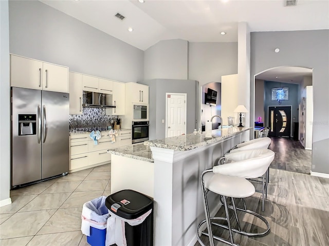 kitchen featuring backsplash, light wood-type flooring, light stone counters, stainless steel appliances, and high vaulted ceiling