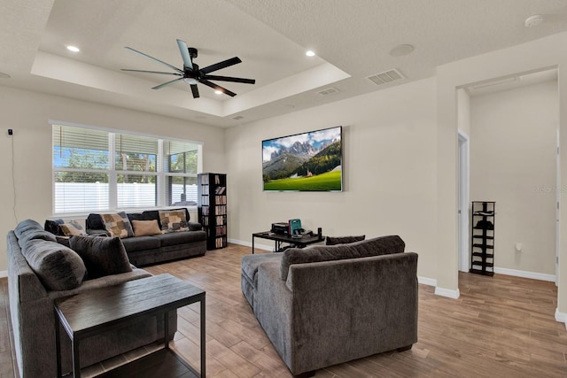 living room with a raised ceiling, a textured ceiling, light hardwood / wood-style flooring, and ceiling fan