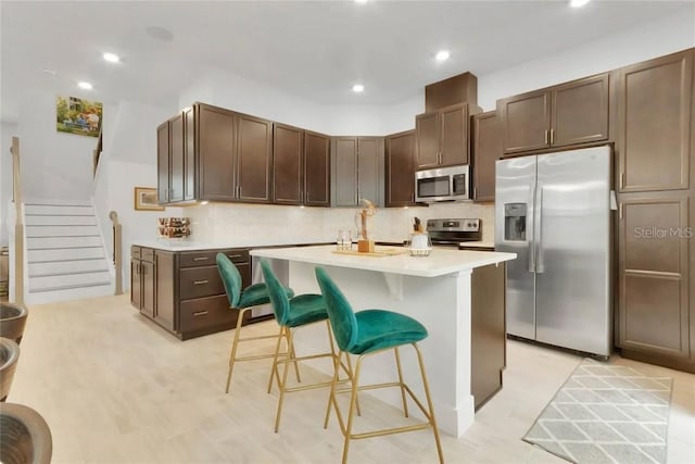 kitchen with dark brown cabinets, appliances with stainless steel finishes, a center island, a kitchen bar, and decorative backsplash