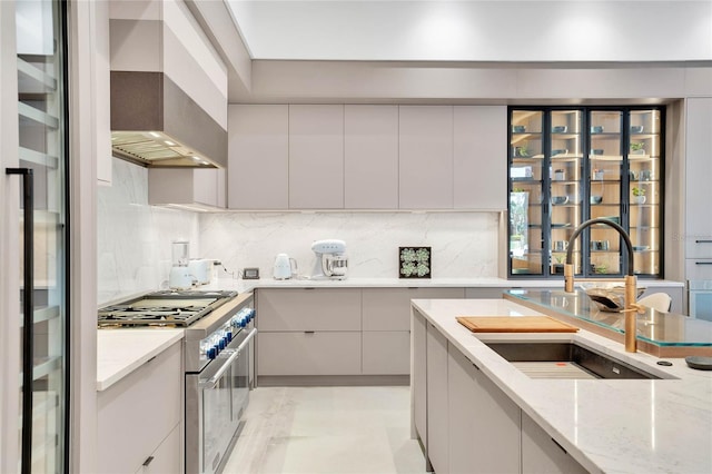 kitchen with light stone countertops, sink, wall chimney exhaust hood, high end stove, and decorative backsplash