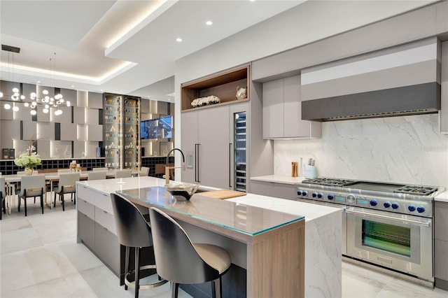 kitchen with decorative backsplash, light stone counters, stainless steel stove, hanging light fixtures, and an island with sink