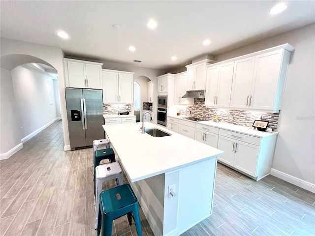 kitchen with arched walkways, an island with sink, appliances with stainless steel finishes, under cabinet range hood, and a sink