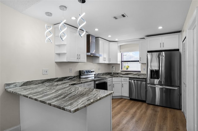 kitchen with kitchen peninsula, hanging light fixtures, wall chimney exhaust hood, appliances with stainless steel finishes, and white cabinets