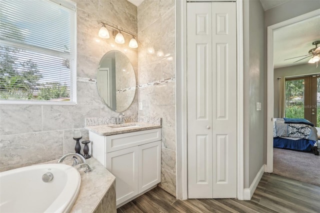 bathroom with a tub to relax in, tile walls, wood-type flooring, ceiling fan, and vanity