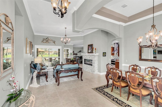 dining space featuring a raised ceiling, an inviting chandelier, a stone fireplace, crown molding, and light tile patterned flooring