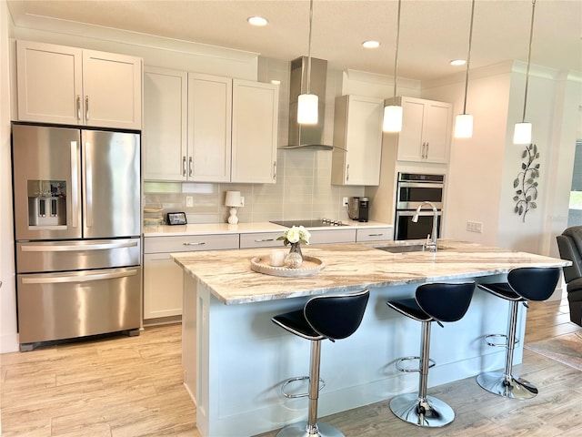 kitchen featuring appliances with stainless steel finishes, light stone counters, an island with sink, and wall chimney range hood