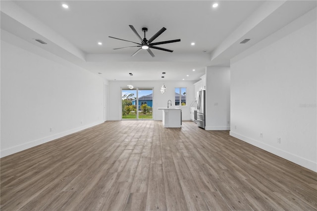 unfurnished living room featuring ceiling fan, a tray ceiling, sink, and light wood-type flooring