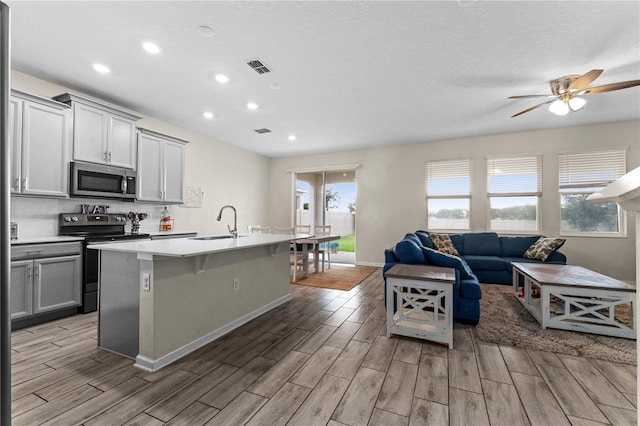 kitchen with gray cabinets, an island with sink, ceiling fan, appliances with stainless steel finishes, and light hardwood / wood-style floors