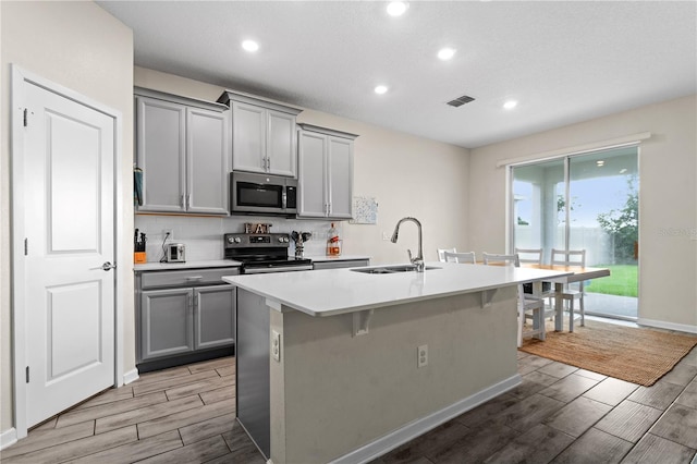 kitchen with light hardwood / wood-style flooring, sink, appliances with stainless steel finishes, a center island with sink, and gray cabinetry