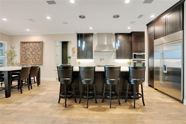 kitchen with dark brown cabinetry, wall chimney exhaust hood, a kitchen island with sink, stainless steel appliances, and light countertops