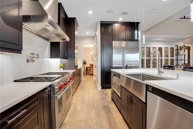 kitchen with premium appliances, a sink, wall chimney range hood, light wood-type flooring, and decorative light fixtures