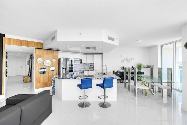 kitchen with a breakfast bar area, appliances with stainless steel finishes, white cabinets, a wall of windows, and dark stone counters