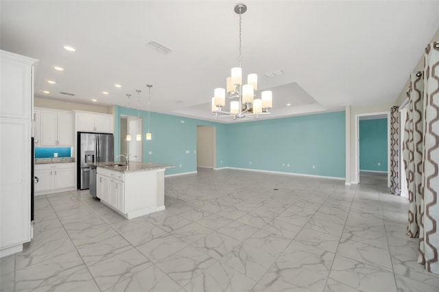 kitchen featuring hanging light fixtures, light stone countertops, white cabinetry, stainless steel fridge, and an island with sink