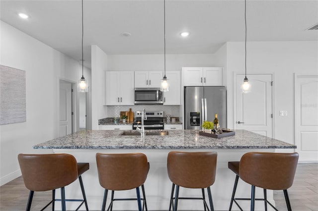 kitchen featuring decorative light fixtures, a center island with sink, backsplash, stainless steel appliances, and light wood-type flooring