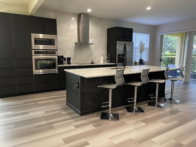 kitchen with a breakfast bar, a center island with sink, dark cabinetry, appliances with stainless steel finishes, and wall chimney exhaust hood