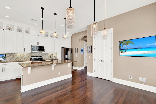 kitchen with dark hardwood / wood-style floors, pendant lighting, appliances with stainless steel finishes, a kitchen island with sink, and dark stone countertops