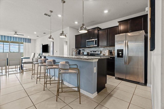 kitchen featuring stainless steel appliances, light stone countertops, an island with sink, decorative backsplash, and decorative light fixtures