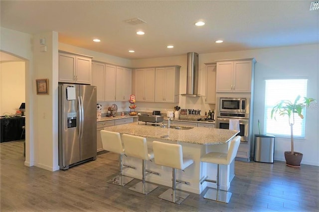 kitchen with an island with sink, appliances with stainless steel finishes, hardwood / wood-style flooring, a breakfast bar area, and wall chimney range hood