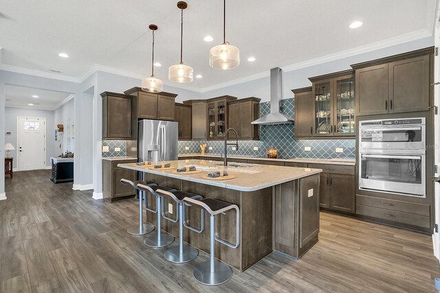 kitchen with hardwood / wood-style floors, appliances with stainless steel finishes, sink, wall chimney range hood, and a kitchen island with sink