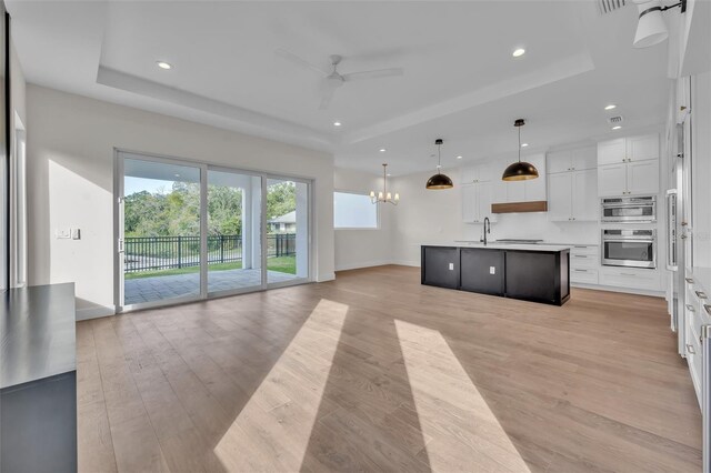 kitchen featuring white cabinets, an island with sink, decorative light fixtures, a tray ceiling, and ceiling fan with notable chandelier