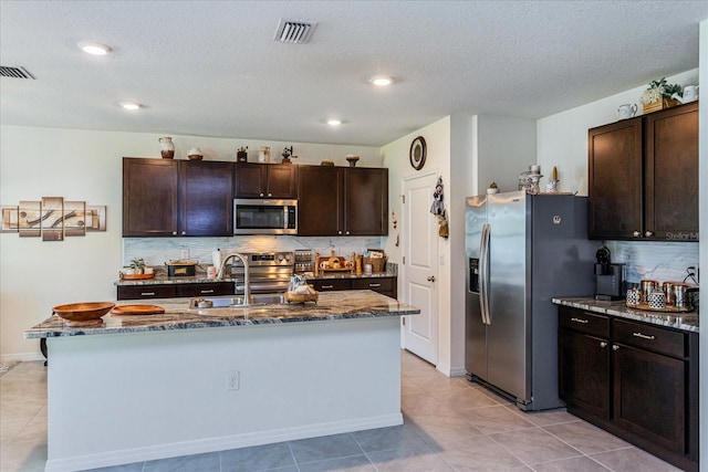 kitchen with stone countertops, an island with sink, sink, decorative backsplash, and appliances with stainless steel finishes