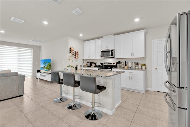 kitchen with light stone countertops, appliances with stainless steel finishes, a breakfast bar area, a center island with sink, and white cabinets