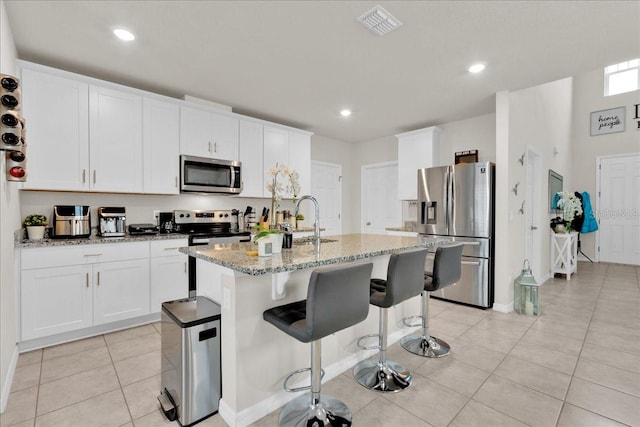 kitchen with a kitchen island with sink, white cabinetry, a kitchen bar, stainless steel appliances, and light stone countertops