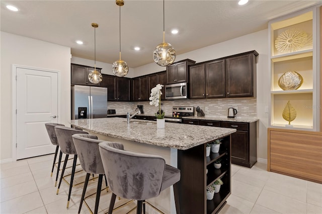 kitchen featuring hanging light fixtures, dark brown cabinets, a center island with sink, stainless steel appliances, and light stone countertops