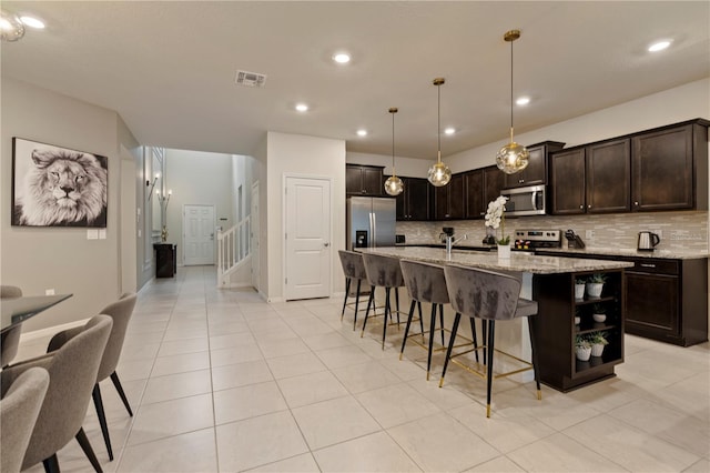 kitchen with appliances with stainless steel finishes, hanging light fixtures, light stone counters, dark brown cabinets, and a center island with sink