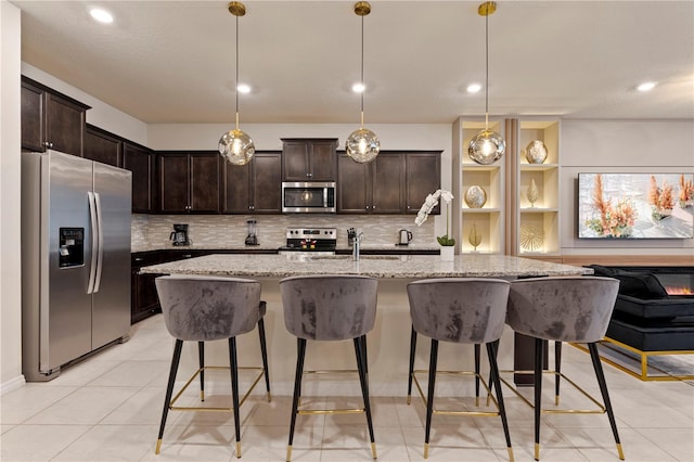 kitchen featuring an island with sink, stainless steel appliances, hanging light fixtures, and light stone countertops