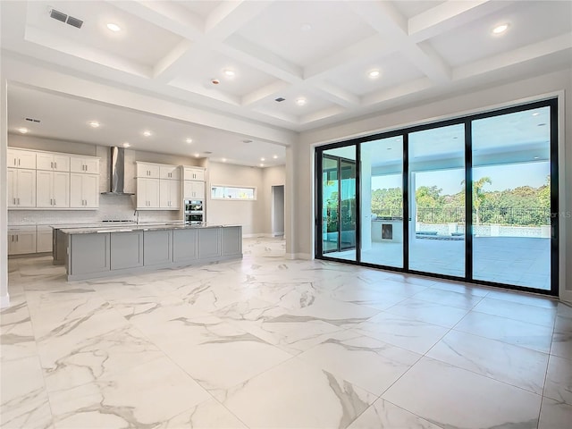 kitchen with decorative backsplash, white cabinets, wall chimney exhaust hood, beamed ceiling, and a center island with sink