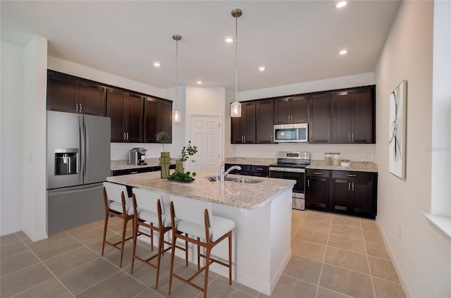 kitchen with decorative light fixtures, appliances with stainless steel finishes, light stone counters, a center island with sink, and dark brown cabinetry