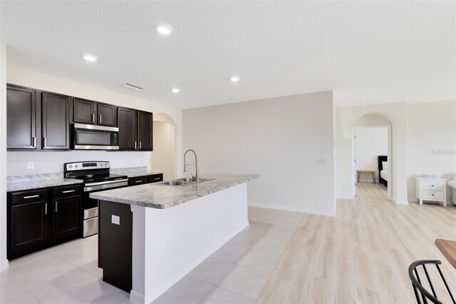 kitchen with a textured ceiling, a kitchen island with sink, stainless steel appliances, sink, and light wood-type flooring