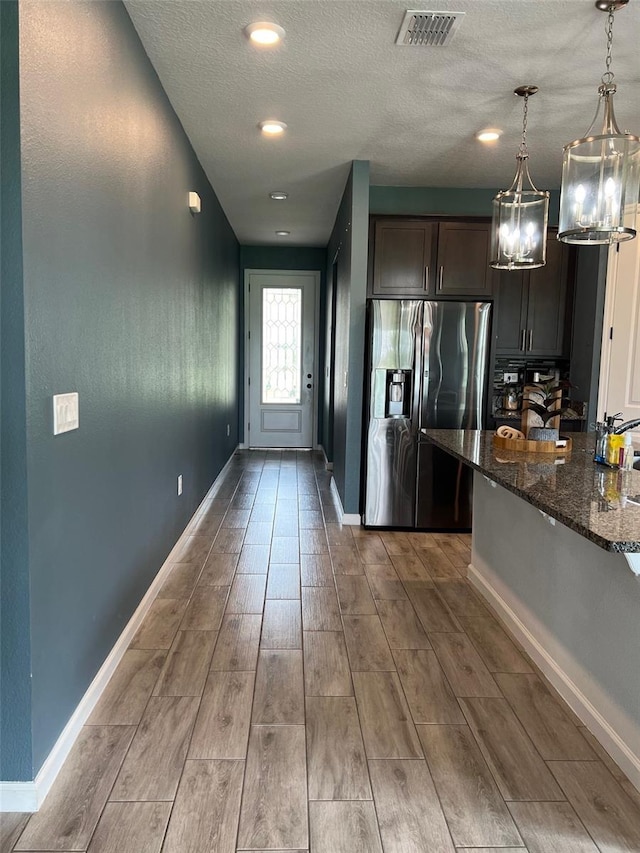 kitchen featuring wood-type flooring, stainless steel refrigerator with ice dispenser, dark brown cabinets, decorative light fixtures, and dark stone countertops