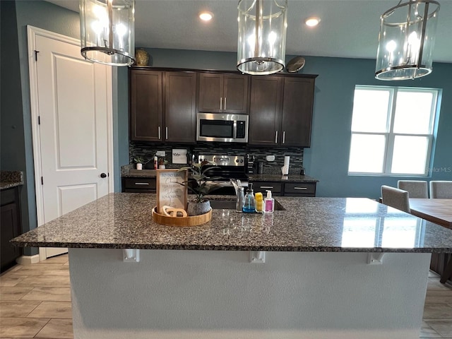 kitchen featuring stainless steel appliances, dark stone counters, light wood-type flooring, pendant lighting, and a kitchen island with sink