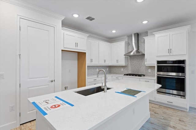 kitchen with wall chimney range hood, an island with sink, white cabinets, and appliances with stainless steel finishes