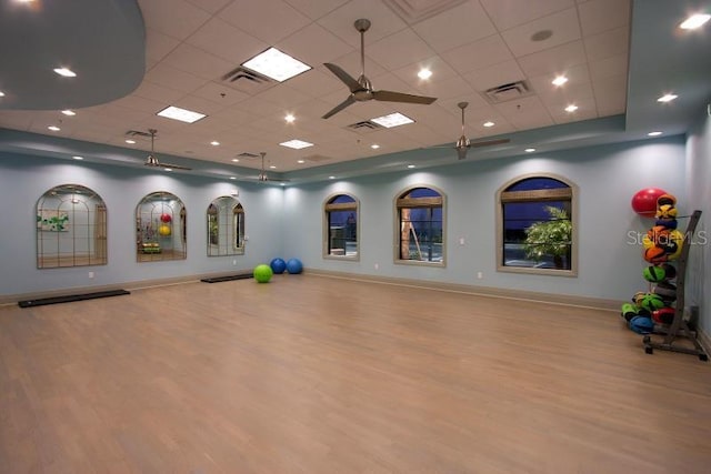 exercise room with a raised ceiling, wood finished floors, visible vents, and ceiling fan