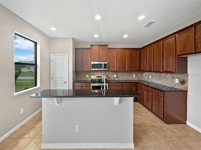 kitchen featuring dark stone counters, backsplash, stainless steel appliances, a kitchen breakfast bar, and a center island with sink