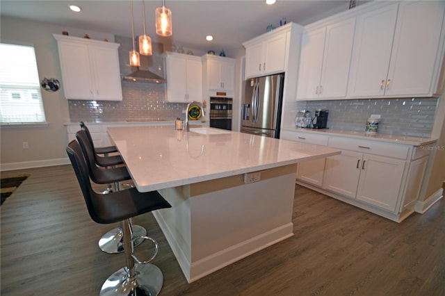 kitchen with appliances with stainless steel finishes, an island with sink, dark wood-type flooring, wall chimney range hood, and white cabinets