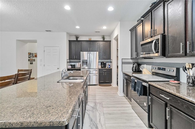 kitchen featuring stone counters, stainless steel appliances, sink, an island with sink, and a textured ceiling