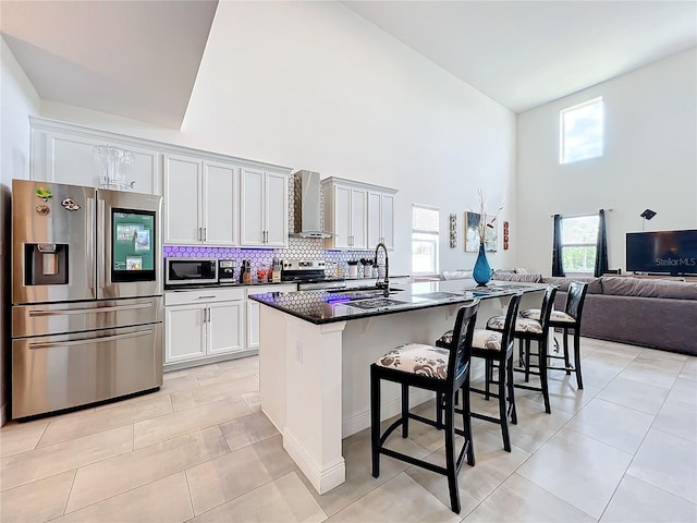 kitchen featuring a kitchen island with sink, a kitchen bar, stainless steel appliances, a towering ceiling, and wall chimney range hood