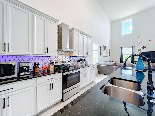 kitchen featuring dark stone countertops, light tile patterned floors, stainless steel appliances, wall chimney exhaust hood, and a high ceiling