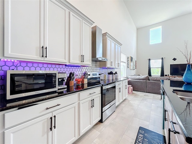 kitchen featuring appliances with stainless steel finishes, high vaulted ceiling, wall chimney range hood, dark stone counters, and light tile patterned flooring