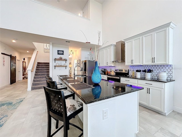 kitchen featuring wall chimney exhaust hood, an island with sink, stainless steel appliances, a barn door, and dark stone countertops