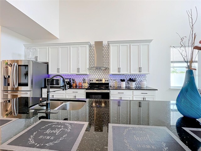 kitchen with dark stone counters, white cabinetry, stainless steel appliances, and wall chimney range hood