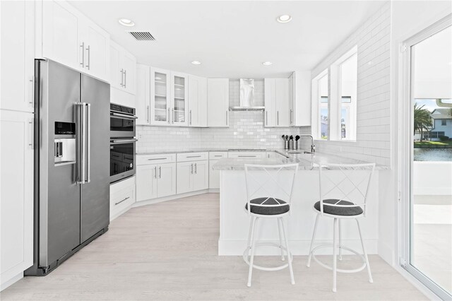 kitchen featuring white cabinets, kitchen peninsula, stainless steel appliances, a kitchen bar, and wall chimney range hood